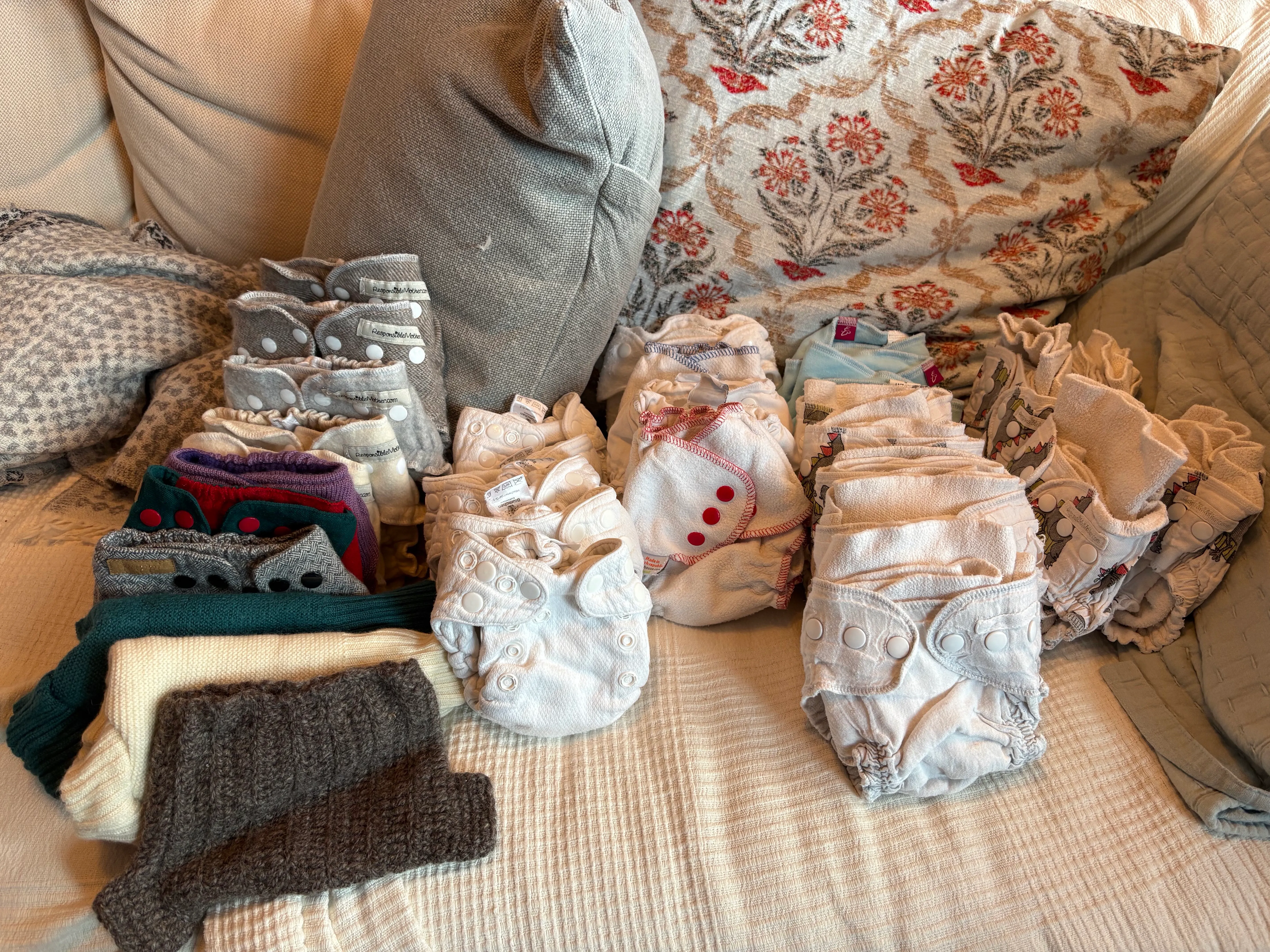 An assortment of folded cloth diapers and knitted wool covers laid out on a bed among cushions