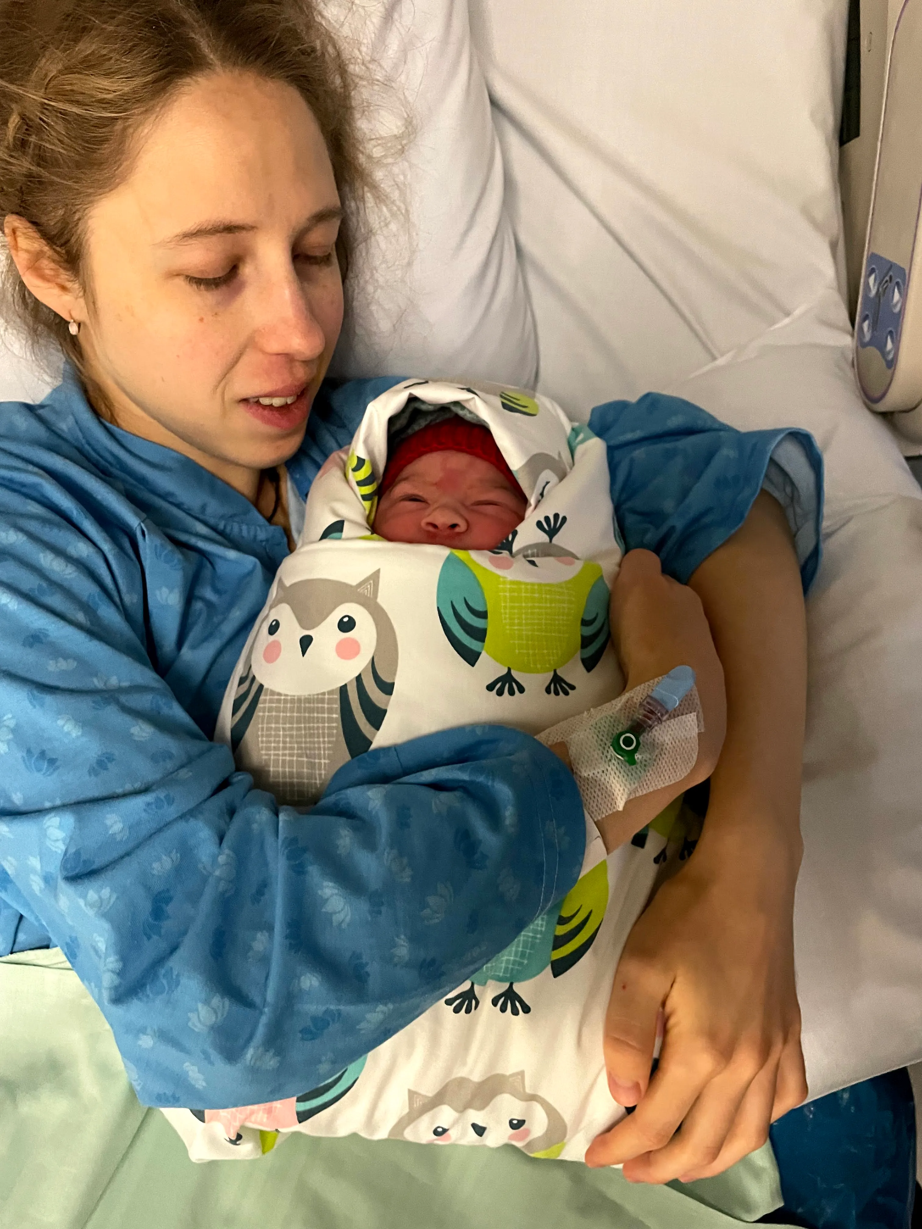 Sara resting in a hospital bed, cradling her newborn daughter who is swaddled in an owl-print blanket and a red knit cap
