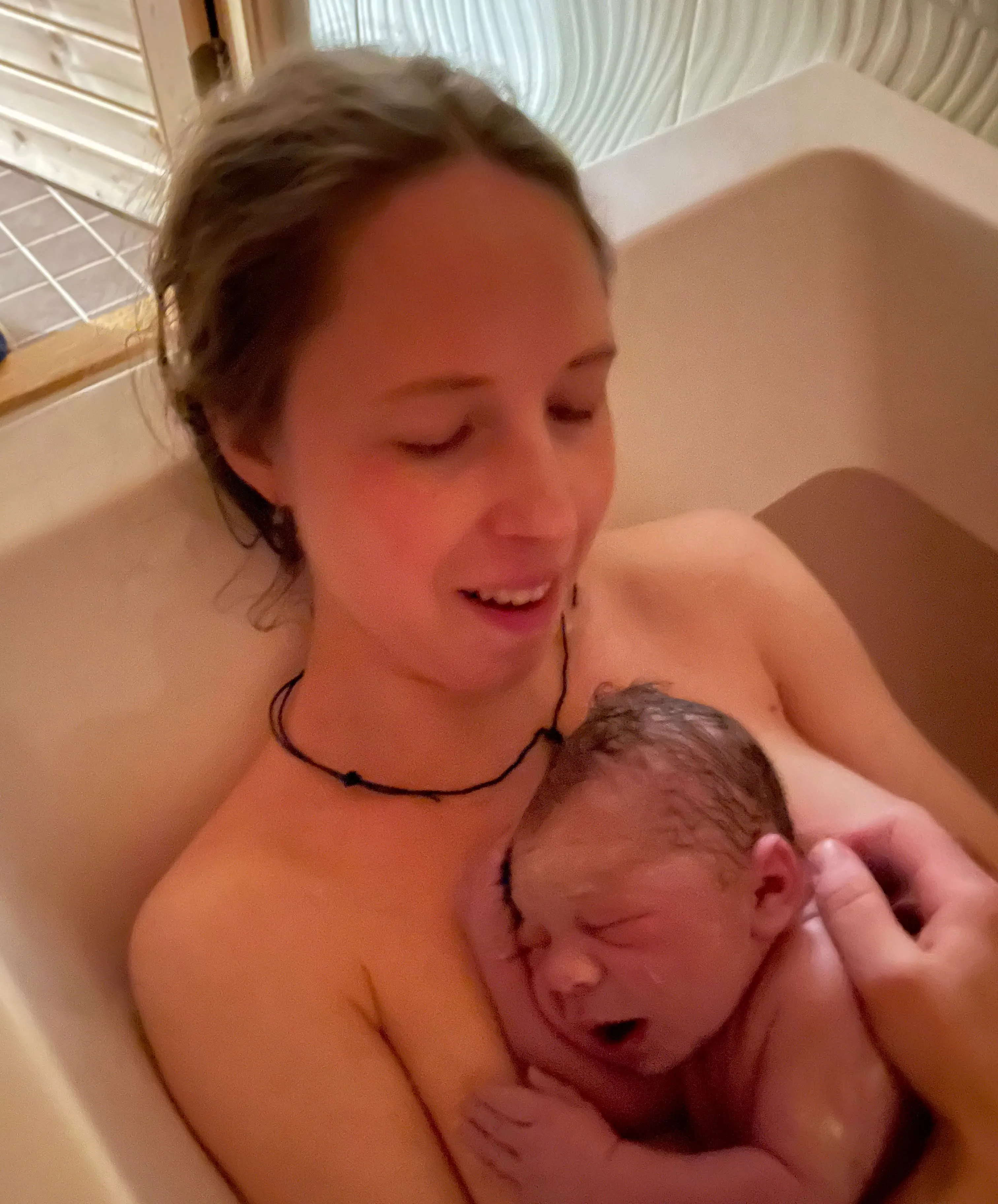 Sara holding her newborn son against her chest in the bathtub just after his birth at home