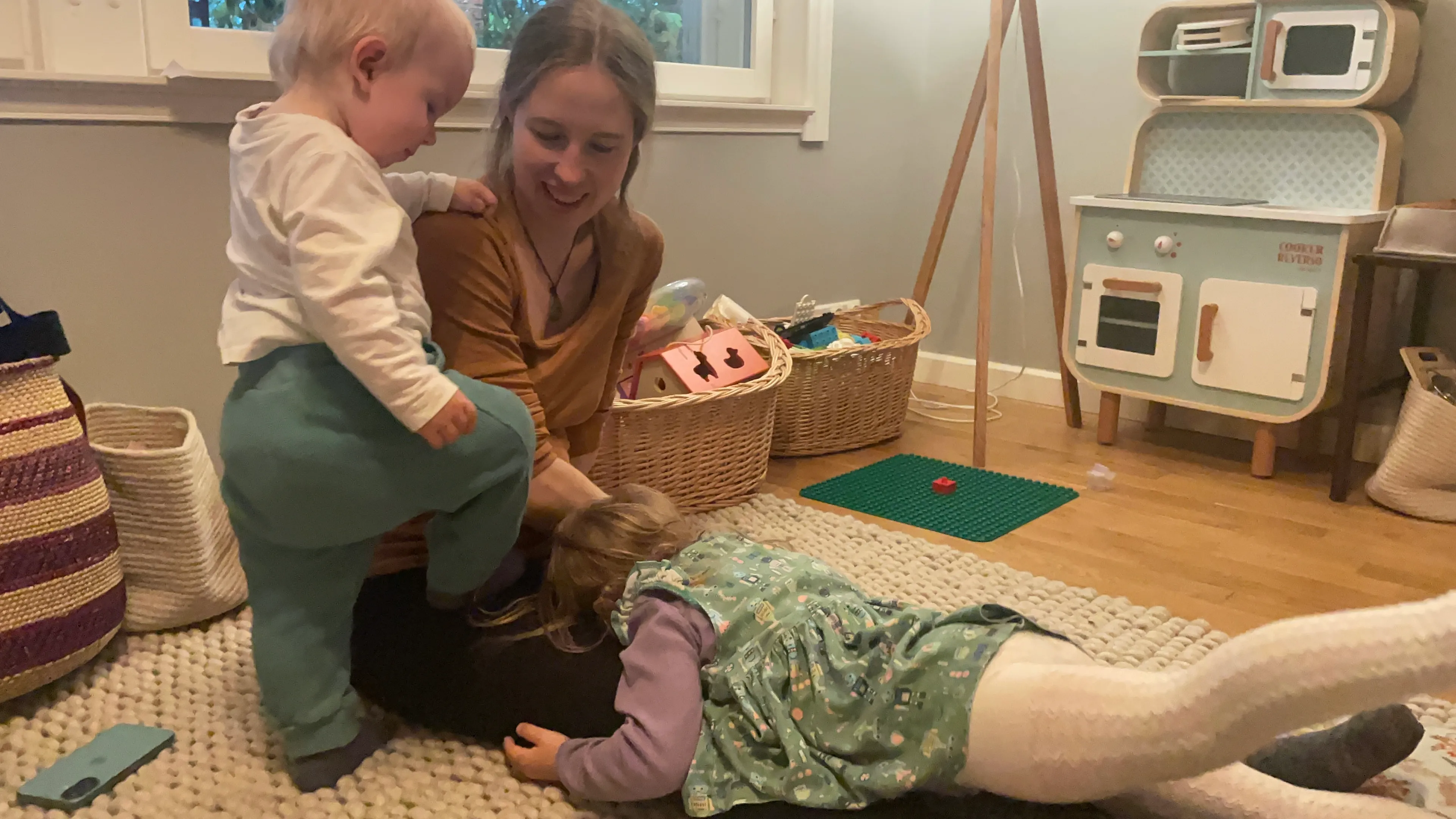 Sara, pregnant, sitting on a playroom floor and smiling as her two young children climb and play on her lap